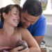 parto-humanizado Mulher com recém-nascido nos braço está de olhos fechados e com um sorriso nos lábios, por trás um homem a sustenta, olhando para o bebê em seus braços. Estão dentro de uma banheira. Uma outra mão tocando o bebê (provavelmente uma parteira) - o parto acabou de acontecer. atendimento humanizado